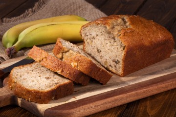 Aufgeschnittenes Bananenbrot mit frischen Bananen im Hintergrund.