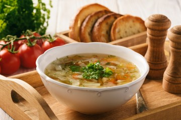 Bild von einer Gemüsesuppe auf einem Holztablett. Im Hintergrund Tomaten und Brot.