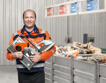 Ein Mitarbeiter des Altstoffsammelzentrums steht vor den Altstoffenstorgungstonnen und hält Elektroschrott in der Hand.