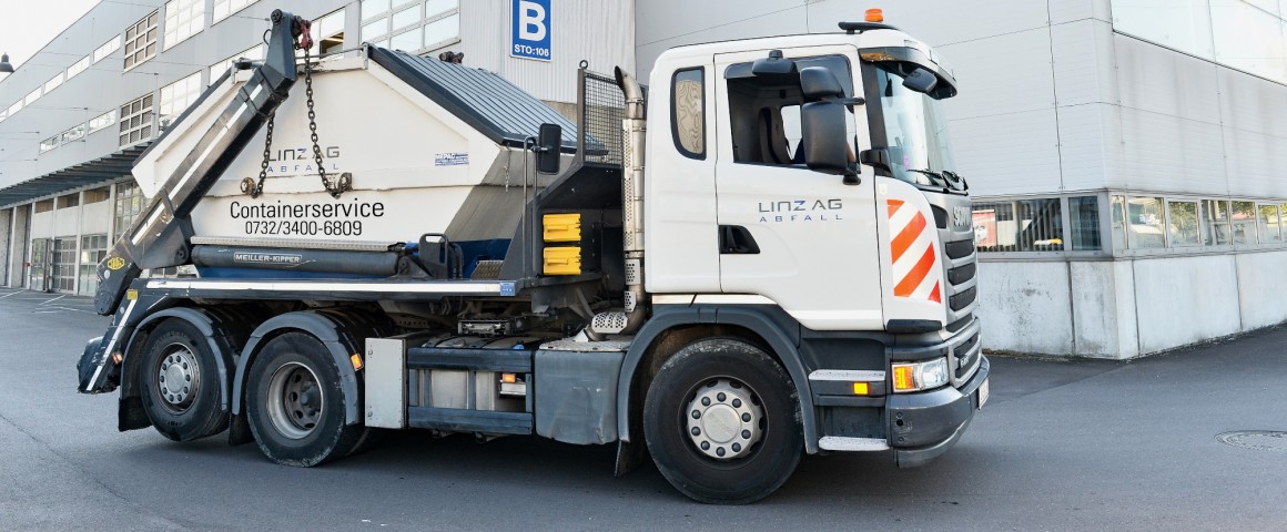 Ein Lastkraftwagen bringt einen Abfallcontainer für den Containerverleih von LINZ AG ABFALL. Im Hintergrund befindet sich ein Gebäude, mit der Aufschrift: LINZ AG Abfall und LINZ AG Abwasser.