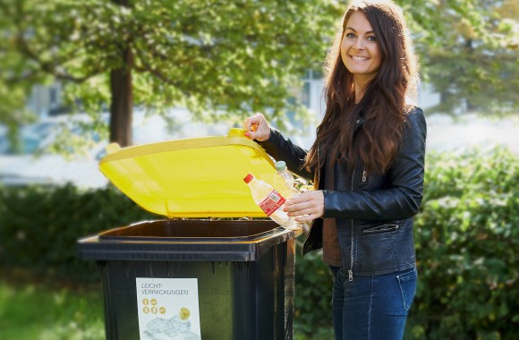 Eine hübsche Lady entsorgt zwei leere Platsikflaschen in der zugehörigen Mülltonne für Leichtverpackungen.