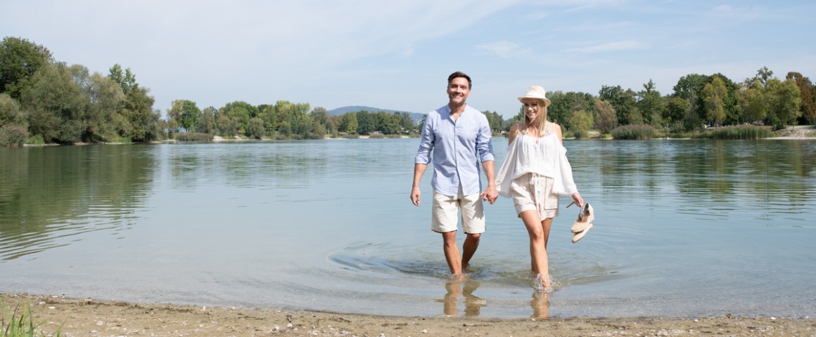 Ein Pärchen hält sich die Hände und spaziert bei schönem Wetter im Wasser am Pichlinger See.