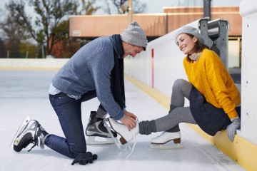Ein Pärchen sitzt am Rand der Eisfläche, der Mann schnürt der Frau die Eislaufschuhe