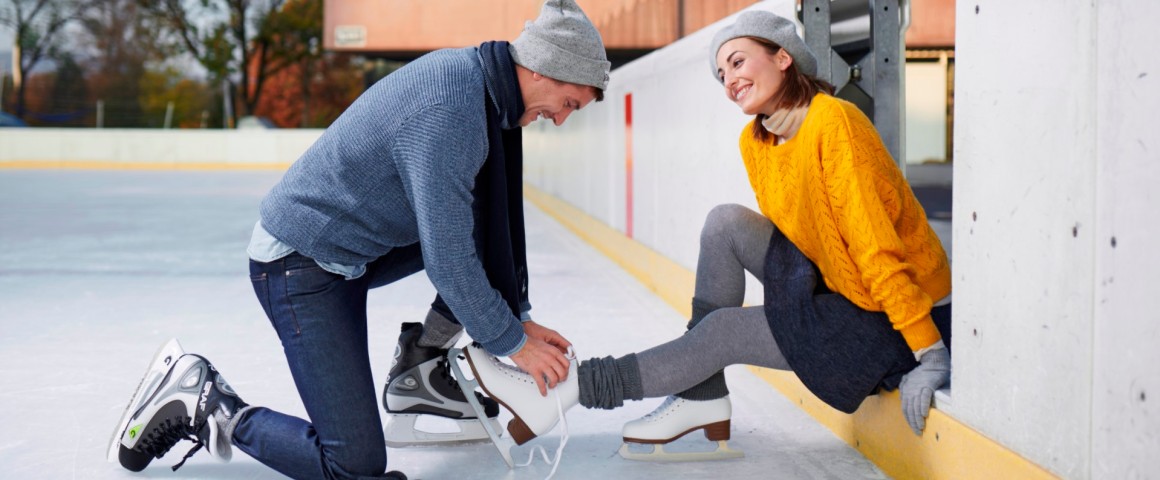 Ein Pärchen sitzt am Rand der Eisfläche, der Mann schnürt der Frau die Eislaufschuhe