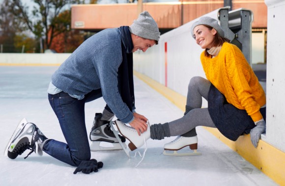 Ein Pärchen sitzt am Rand der Eisfläche, der Mann schnürt der Frau die Eislaufschuhe