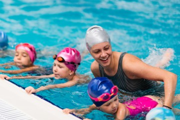 3 Kinder halten sich am Beckenrand fest und lernen schwimmen, die Schwimmlehrerin hilft ihnen die Position zu halten