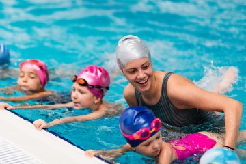 3 Kinder halten sich am Beckenrand fest und lernen schwimmen, die Schwimmlehrerin hilft ihnen die Position zu halten