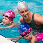 3 Kinder halten sich am Beckenrand fest und lernen schwimmen, die Schwimmlehrerin hilft ihnen die Position zu halten