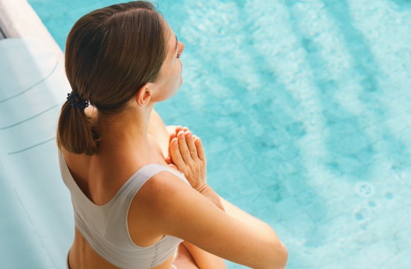 Eine Frau sitzt im Schwimmbecken am Beckenrand und macht eine Yogapose.