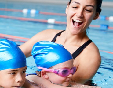 3 Kinder halten sich an ihrem Schwimmbrett fest und lernen schwimmen, die Schwimmlehrerin hilft ihnen die Position zu halten