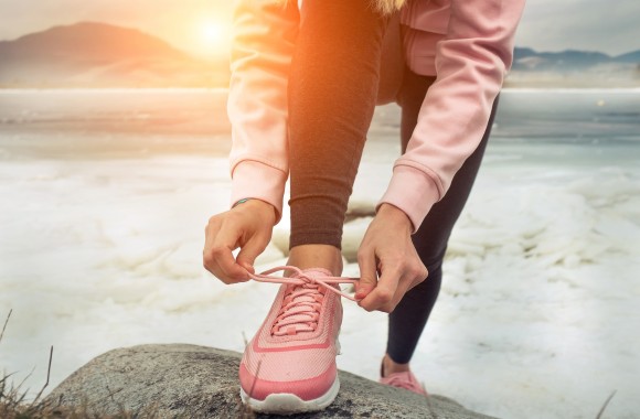 Frau steht im Trainingsoutfi mit dem Rücken zum See und bindet sich die Sportschuhe