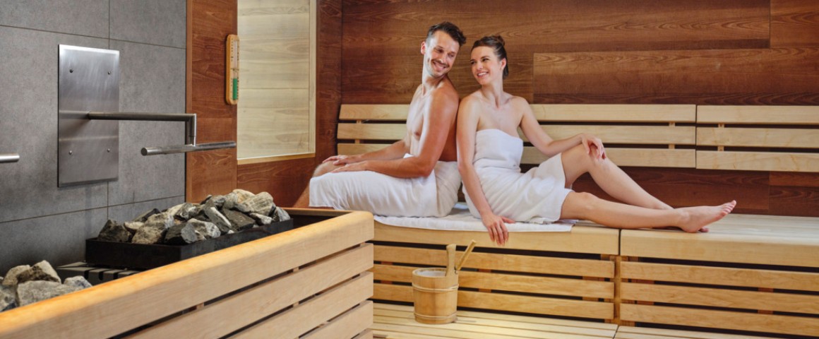 Ein junges Pärchen sitzt Rücken an Rücken in der Sauna und lächtelt sich an.