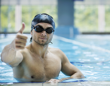 Mann mit Taucherbrille und Badehaube, der den Daumen hochhält, im Schwimmbecken
