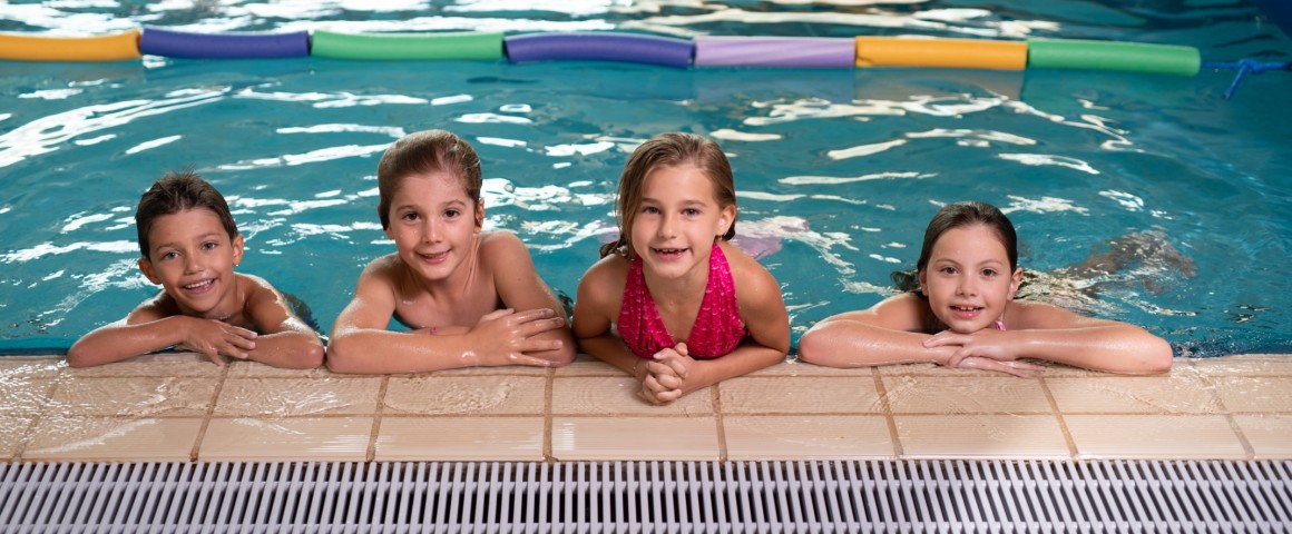 4 Kinder (2 Jungen und 2 Mädchen) halten sich am Beckenrand fest und lächeln in die Kamera.