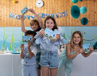 4 Kinder (2 Jungen und 2 Mädchen) stehen nebeneinander und halten Einladungskarten für einen Kindergeburtstag in Linz AG Bädern in die Kamera. Dahinter befindet sich ein Geburtstagstisch und eine Happy Birthday Girlande.