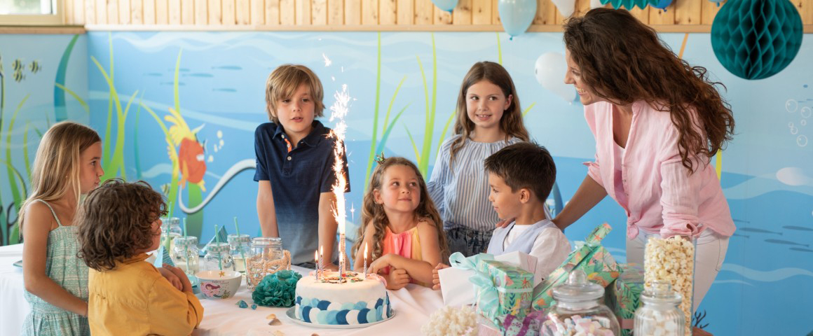 Bild von einem Kindergeburtstag. 6 Kinder (3 Jungen und 3 Mädchen) mit einer Mutter sind um einen Tisch versammelt. Auf dem Tisch befinden sich Süßigkeiten und eine Torte mit einer Spritzkerze darauf.