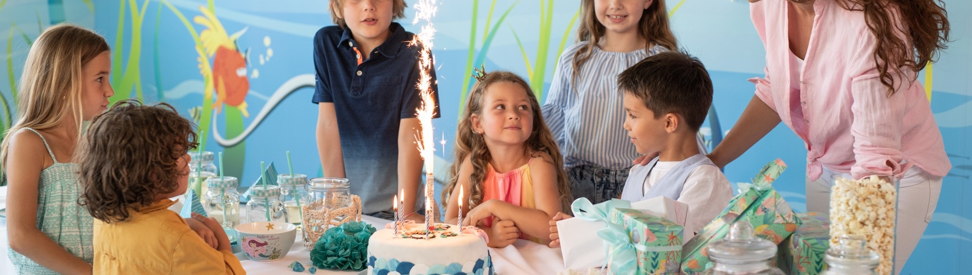 Bild von einem Kindergeburtstag. 6 Kinder (3 Jungen und 3 Mädchen) mit einer Mutter sind um einen Tisch versammelt. Auf dem Tisch befinden sich Süßigkeiten und eine Torte mit einer Spritzkerze darauf.