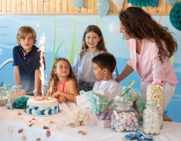 Bild von einem Kindergeburtstag. 6 Kinder (3 Jungen und 3 Mädchen) mit einer Mutter sind um einen Tisch versammelt. Auf dem Tisch befinden sich Süßigkeiten und eine Torte mit einer Spritzkerze darauf.