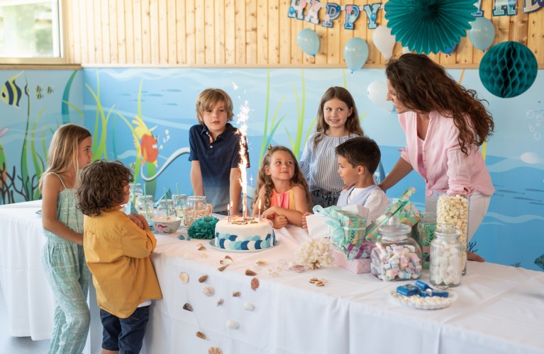 Bild von einem Kindergeburtstag. 6 Kinder (3 Jungen und 3 Mädchen) mit einer Mutter sind um einen Tisch versammelt. Auf dem Tisch befinden sich Süßigkeiten und eine Torte mit einer Spritzkerze darauf.