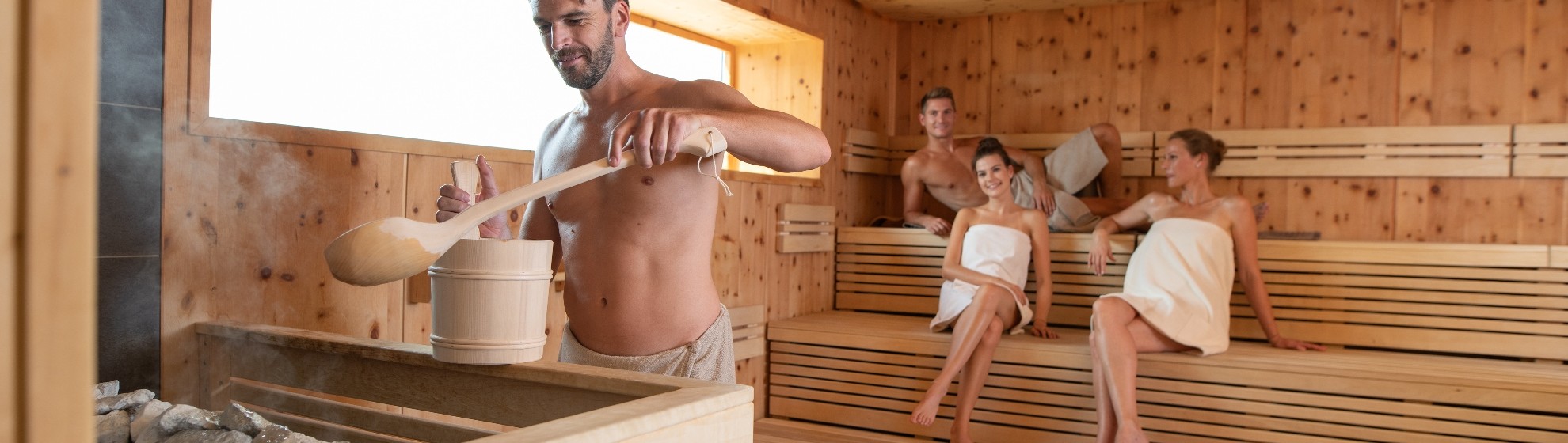Bild von Sauna, in der gerade ein Aufguss von einem Mann gemacht wird. Im Hintergrund sitzen zwei Frauen und ein Mann.