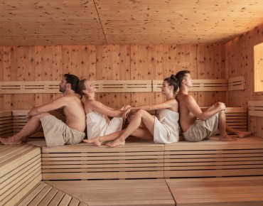 Aufnahme in Sauna. Mann sitzt Rücken an Rücken mit Frau, gegenüber sitzt Frau Rücken an Rücken mit Mann.