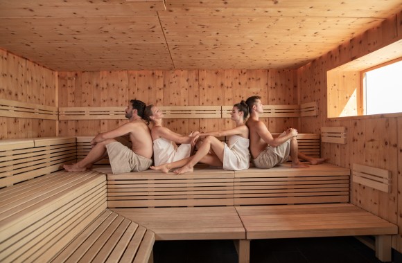 Aufnahme in Sauna. Mann sitzt Rücken an Rücken mit Frau, gegenüber sitzt Frau Rücken an Rücken mit Mann.