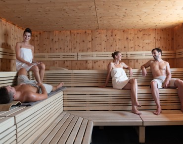 Bild in Sauna, in der sich 2 Pärchen befinden. 1 Mann liegt, alle anderen sitzen.