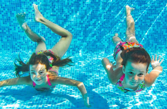 Unterwasseraufnahme von zwei jungen Mädchen, welche in die Kamera lächeln.