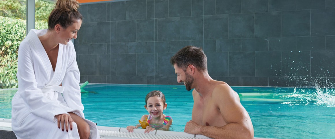 Eine Frau sitzt mit  Bademantel am Beckenrand eines Schwimmbeckens und ein Kind und ein Mann befinden sich mit Badebekleidung im Wasser.
