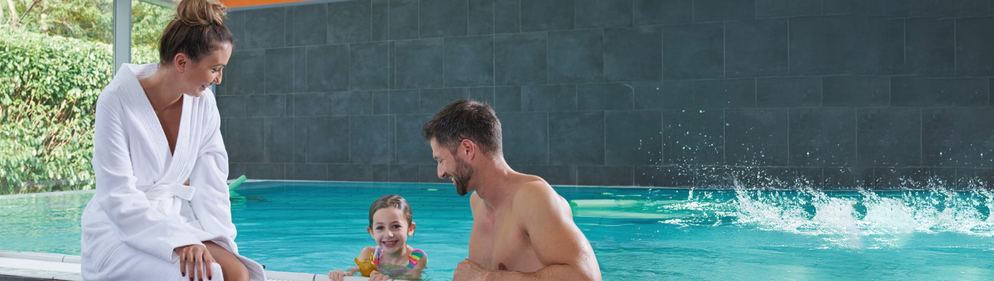 Eine Frau sitzt mit  Bademantel am Beckenrand eines Schwimmbeckens und ein Kind und ein Mann befinden sich mit Badebekleidung im Wasser.