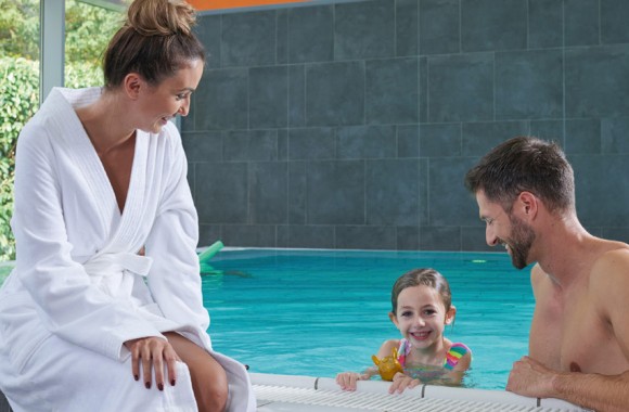 Eine Frau sitzt mit  Bademantel am Beckenrand eines Schwimmbeckens und ein Kind und ein Mann befinden sich mit Badebekleidung im Wasser.