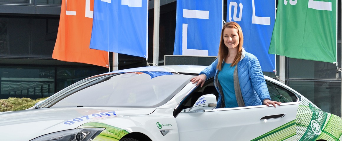 Eine Frau posiert mit einem E-Auto mit LINZ AG-Branding, einem Tesla, vor der LINZ AG