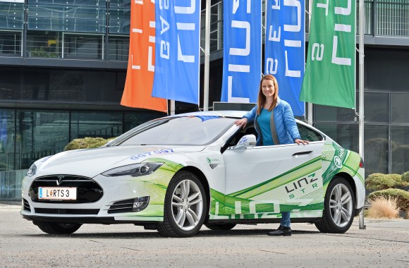 Eine Frau posiert mit einem E-Auto mit LINZ AG-Branding, einem Tesla, vor der LINZ AG