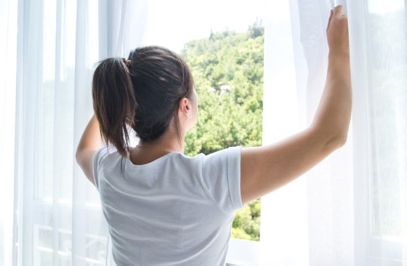 Junge Frau steht vor Fenster mit schöner Aussicht und schiebt weiße Vorhänge zur Seite