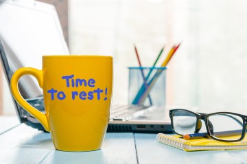 Schreibtisch auf dem eine gelbe Tasse mit der Aufschrift "Time to rest!", Brille, Notizheft, Stift, Stiftehalter und einem Laptop.