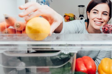 Eine Frau steht vor dem geöffneten Kühlschrank und greift lachend zu einer Zitrone.