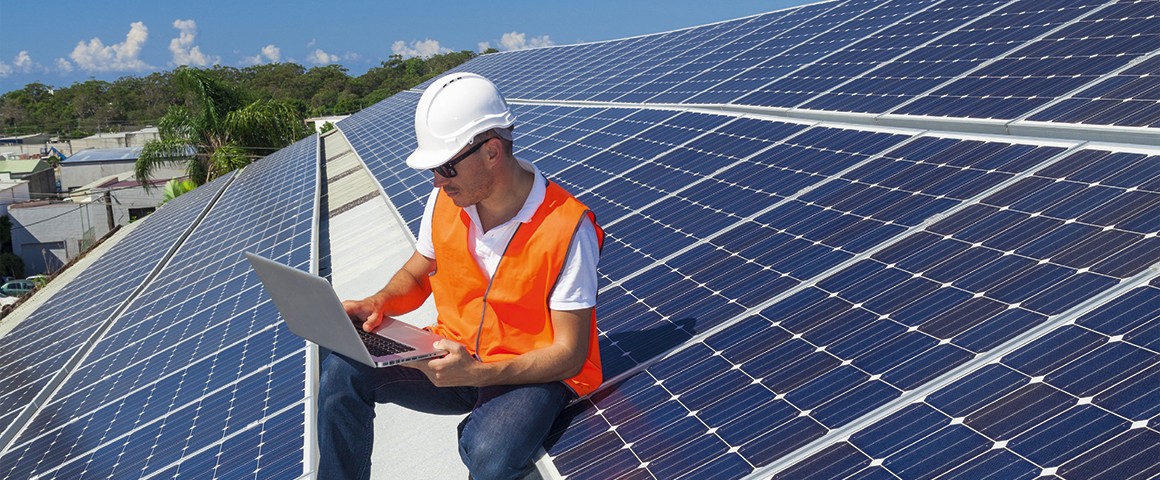 Mann sitzt auf einer Solaranlage am Dach eines Gebäudes und schaut in den Laptop