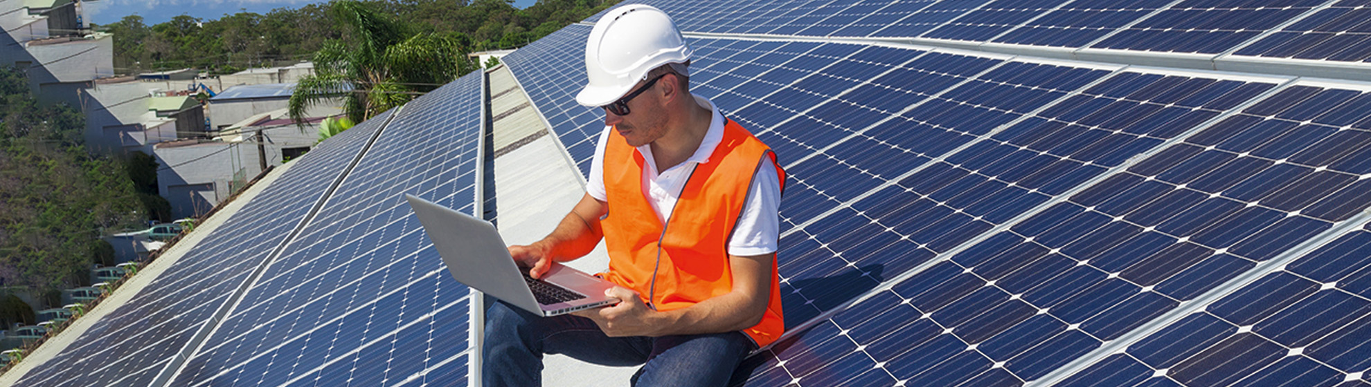 Mann sitzt auf einer Solaranlage am Dach eines Gebäudes und schaut in den Laptop