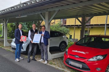 Der Geschäftsführer der Volkshilfe Oberösterreich und zwei Personen der Linz AG stehen beim PV-Carport der Volkshilfe und halten eine Urkunde in die Kamera