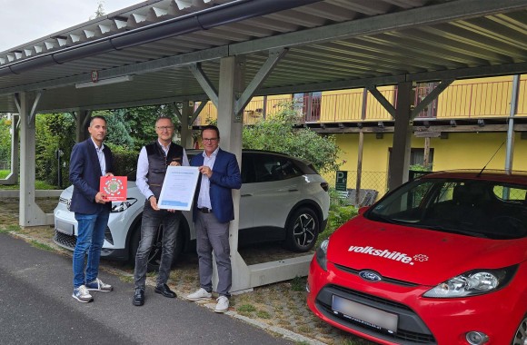Der Geschäftsführer der Volkshilfe Oberösterreich und zwei Personen der Linz AG stehen beim PV-Carport der Volkshilfe und halten eine Urkunde in die Kamera