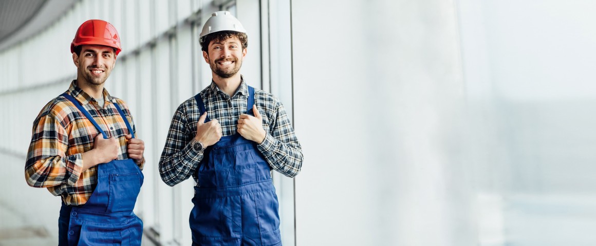 Zwei Arbeiter im Blaumann und Helm stehen in einem Gebäude 