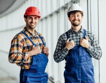 Zwei Arbeiter im Blaumann und Helm stehen in einem Gebäude 