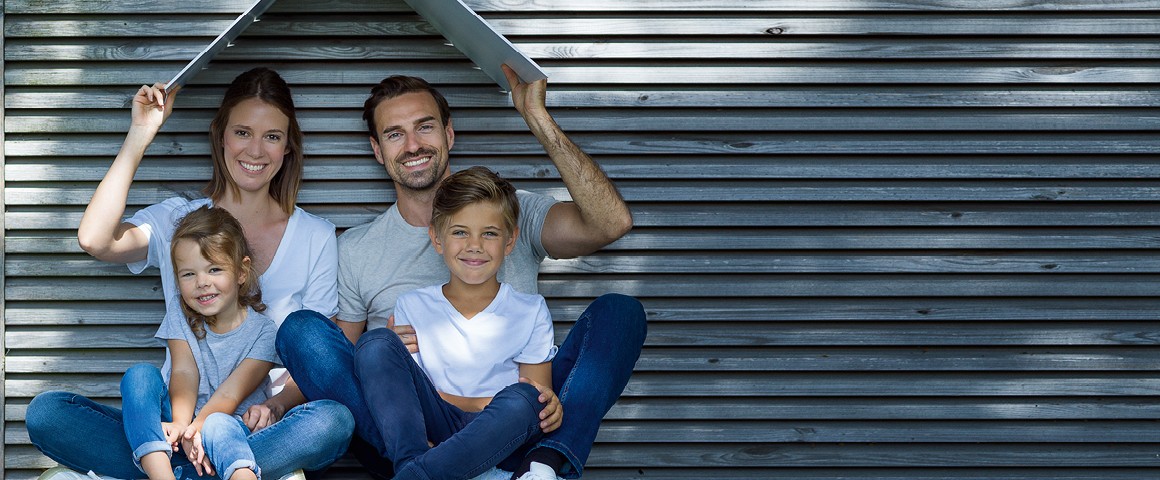 Familie (1 Junge und 1 Mädchen sitzen auf dem Schoß ihrer Eltern) sitzt vor einer Hausmauer und lacht