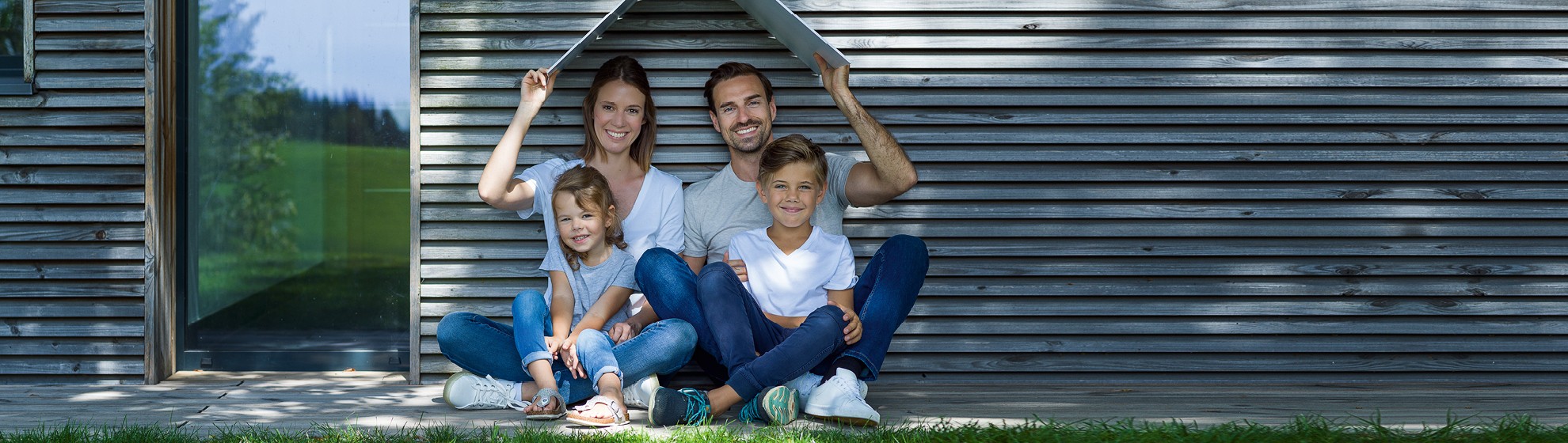 Familie (1 Junge und 1 Mädchen sitzen auf dem Schoß ihrer Eltern) sitzt vor einer Hausmauer und lacht