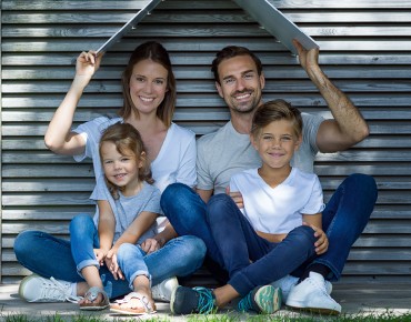 Familie (1 Junge und 1 Mädchen sitzen auf dem Schoß ihrer Eltern) sitzt vor einer Hausmauer und lacht