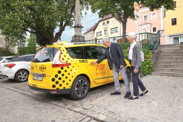 Zwei Senioren (ein Mann und eine Frau) steigen in ein gelbes AST Auto ein.