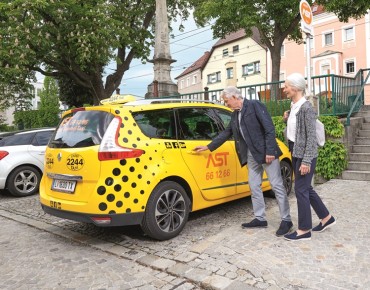 Zwei Senioren (ein Mann und eine Frau) steigen in ein gelbes AST Auto ein.