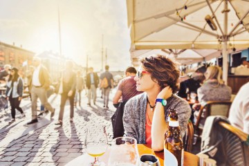 Junge Frau sitzt in einem Straßencafé bei einem Getränk und beobachtet die Umgebung