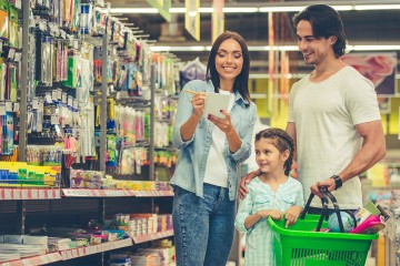 Eine Familie (Vater, Mutter, Kind) in einem Geschäft vor einem Regal mit Schularitkeln. Die Mutter hält einen Zettel in der Hand und hakt einen Eintrag ab.