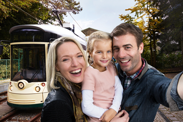Zwei Eltern und ihre Tochter lächeln fröhlich in die Kamera. Im Hintergrund ist die Pöstlingbergbahn zu sehen.